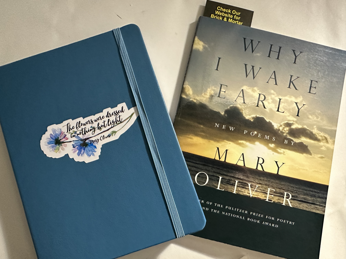 Picture of a blue notebook with a Mary Oliver quote that says, "The flowers were dressed in nothing but light." The notebook is next to a copy of the Mary Oliver poetry collection, WHY I WAKE EARLY.